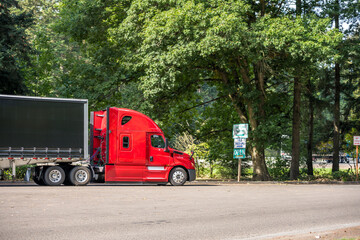 Fototapeta na wymiar Bright red big rig semi truck with covered black semi trailer standing on the rest area parking lot parallel to the highway