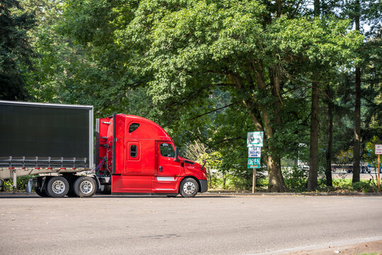 Bright red big rig semi truck with covered black semi trailer standing on the rest area parking lot parallel to the highway