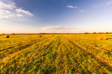 Scenic view at picturesque burning sunset in a green shiny field with hay stacks, bright cloudy sky , trees and golden sun rays, summer valley landscape