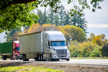 Convoy of two different big rigs semi trucks with dry van and container semi trailers running on the summer road with green trees