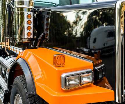 Fragment Of Front Part Of Orange And Black Big Rig Semi Truck Tractor With Shiny Paint And Chrome Accessories