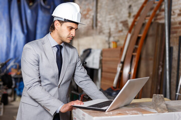 Confident male architect in suit and helmet controlling process of building using laptop