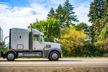 Profile of industrial big rig semi truck tractor with semi trailer driving on the flat highway road with trees on the side