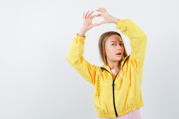 Fototapeta na wymiar blonde girl in pink t-shirt and yellow jacket showing love gesture with hands above head and looking alluring , front view.