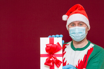 Handsome male wears Santa hat medical mask gloves holds gift box showing on camera red studio background.