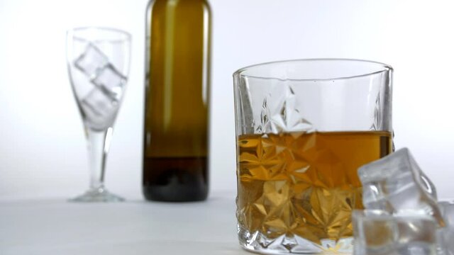Crystal clear whiskey glass kept on a table with ice cubes placed near it. Pan shot of an empty glass  alcohol bottle  and filled whiskey glass against a white background