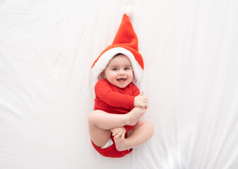 funny baby girl 6 month in red bodysuit and Santa hat lying on white bedding. minimalist. top view.
