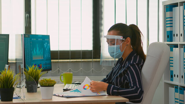 Manager woman wearing face protection against coronavirus typing on computer analysing new statistics data working in new normal financial office. Freelancer in company respecting social distance.