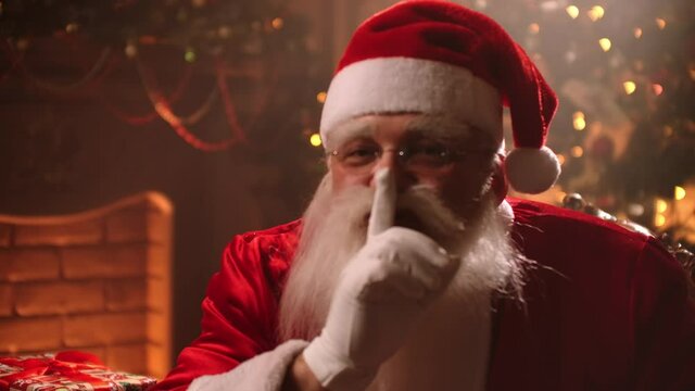 actor is playing role of Santa Claus, holding finger near lips for silence at Christmas decorated room, hush sign at gesturing