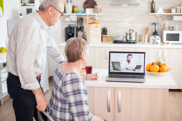 Doctor on telemedicine for senior couple. Video conference with doctor using laptop in kitchen. Online health consultation for elderly people drugs ilness advice on symptoms, physician telemedicine