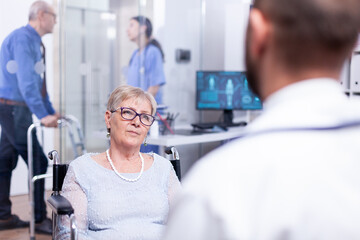 Fototapeta premium Handicapped old woman sitting in wheelchair during medical examination with doctor and listening diagnosis. Recovery treatment, help for disabled people. Elderly assistance