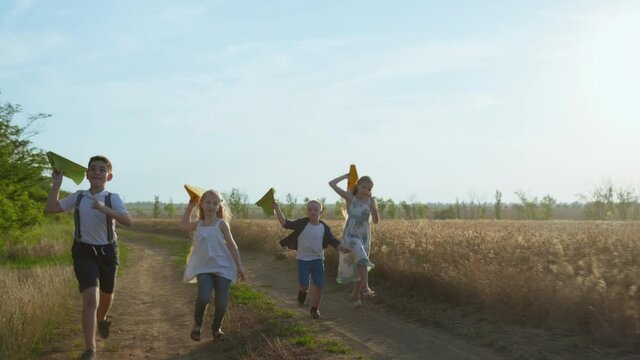 group of friends, boys and girls running along a dusty country road and launch paper planes into the air near the golden wheat field