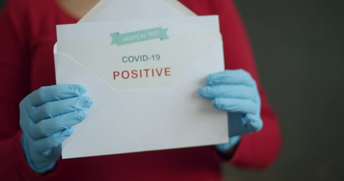 Woman Holding Positive Antigen Detection Test for Covid-19. Push In of Person Wearing Face Mask and Blue Surgical Gloves Showing Open Envelope with Positive Coronavirus Test Results into Camera.