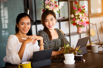 Two beautiful Asian women Sitting at work online or studying during the corona virus with a tablet in the morning, the sun warms happily.