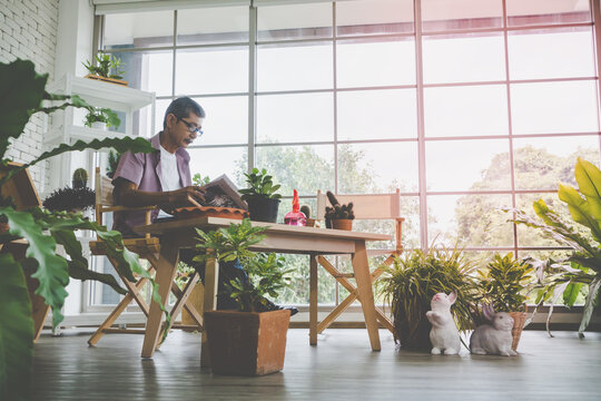 Asian Senoir Man Is Relaxing While Reading Newspaper In His Home House Plant Garden.