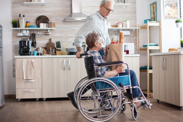 Senior man taking grocery paper bag from handicapped wife in wheelchair. Mature people with fresh...