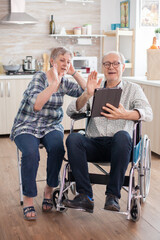Cheerful senior woman waving on video conference in the kitchen. Disabled senior man in wheelchair and his wife having a video conference on tablet pc in kitchen. Paralyzed old man and his wife having