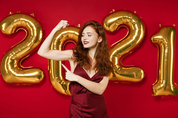 Strong young redhead woman in elegant dress point index finger on biceps muscles isolated on red background, golden numbers air balloons studio. Happy New Year 2021 celebration holiday party concept.