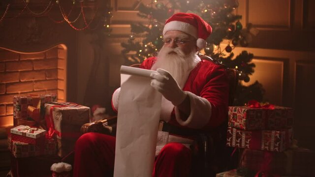Santa Claus is reading list of good kids and checking gifts for Christmas, sitting in his decorated room at festive evening