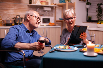 Elderly woman browsing on tablet pc and disabled husband in wheelchair holding wine glass. Imobilized handicapped senior husband browsing on phone enjoying the festive male, drinking a glass of red