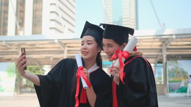 Couple of Asian beautiful women with graduation gown use mobile phone to selfie together in big city. Concept of celebration and congratulation after success of education in high level.