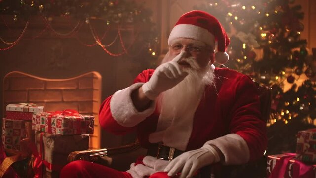 kind funny Santa Claus is sitting alone at room with Christmas decorations at New year Eve, showing silence sign by finger and mouth