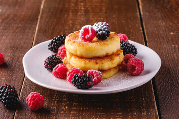 Cottage cheese pancakes and powdered sugar, curd fritters dessert with raspberry and blackberry berries in plate on dark brown wooden background, angle view macro