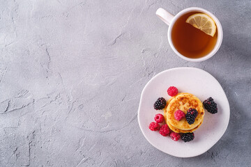 Cottage cheese pancakes, curd fritters dessert with raspberry and blackberry berries in plate near to hot tea cup with lemon slice on stone concrete background, top view copy space