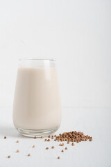 Gluten-free and lactose-free healthy buckwheat milk full of vitamins, minerals and antioxidants served in tall drinking glass with grain ingredient on white wooden background at breakfast. Vertical