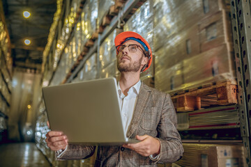Warehouse manager in orange helmet holding a laptop and inspecting a warehouse