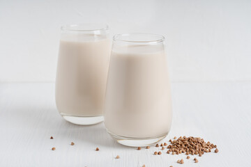 Gluten-free and lactose-free healthy buckwheat milk full of vitamins, minerals and antioxidants served in two tall drinking glasses with raw grain ingredient on white wooden background. Horizontal