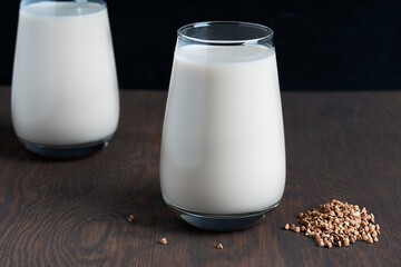 Gluten-free and lactose-free healthy vegan buckwheat milk full of vitamins, minerals and antioxidants served in two drinking glasses with raw seeds ingredient on dark wooden background. Horizontal