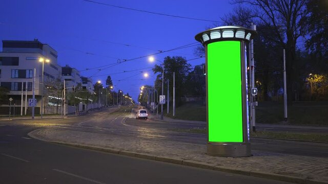 City light vitrine with empty green screen on roadside against cars driving along empty night street. 
