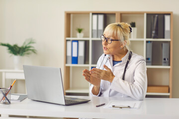 Senior female family doctor uses a laptop to conduct an online consultation with her patient.