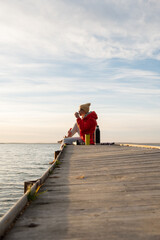 A woman in a red jacket and hat drinks tea on a wooden pier by the lake looking into the distance at dawn