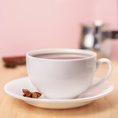 White ceramic cup of coffee on plate with half star anise, cinnamon sticks and turka behind pink background