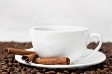 White cup of black coffee with plate and three cinnamon sticks on beans and light background