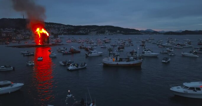 Beautiful, cinematic, approaching, very low altitude aerial time lapse that reveals the biggest bonfire event on Earth and the small boats that are gathering around it near Alesund, Norway at dusk.