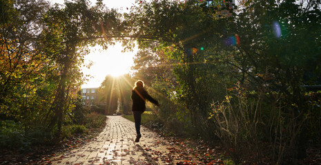 an unidentifiable girl is jumping joyfully through an arch of greenery into the late afternoon...