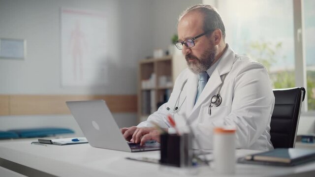 Doctor's Office: Hispanic Physician Sitting at His Desk Working on Personal Computer. Medical Health Care Specialist Filling Prescription Forms, Checking Analysis Test Results. Arc Side View Shot