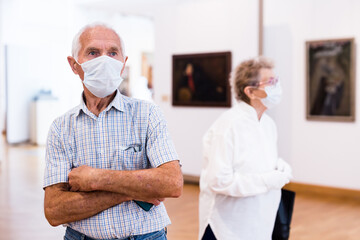 mature man in mask protecting against covid examines paintings on display in hall of art museum