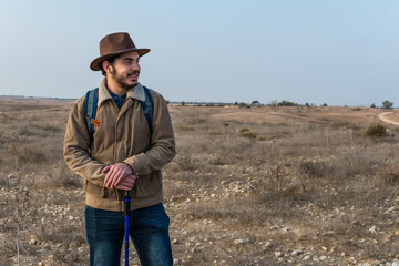 Hiker young man with beard, wearing brown jacket, backpack and a hipster hat. Outdoors background.