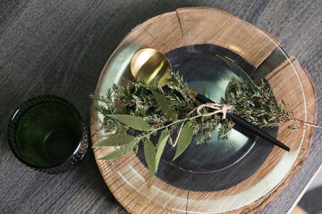 table setting decorated for the new year with cutlery, blue tablecloth, stand made of wood, transparent plate, glass, thuja branch, golden spoon 