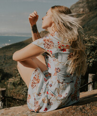 A woman sitting on a boulder looking out at the view in front of her, feeling the wind blow through her long blond hair.