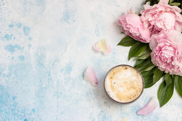 Morning cup of coffee or cappuccino and pink peonies on blue table. Top view. Creative breakfast for Woman or Mother day.