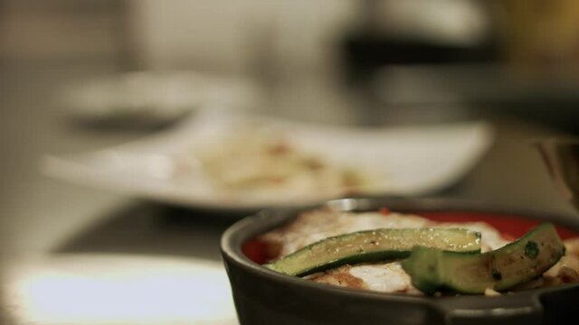 Chef putting grilled Courgette slices on grilled vegetables with fried egg and more vegetables on a side dish behind. Racking Focus.