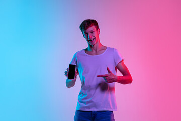 Fototapeta na wymiar Pointing on cellphone. Young caucasian man's portrait on gradient blue-pink studio background in neon light. Concept of youth, human emotions, facial expression, sales, ad. Beautiful model in casual.