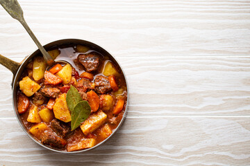 Delicious stew with meat, potatoes, carrot and gravy in rustic copper pot on white wooden background from above. Traditional winter and autumn dish beef and vegetables ragout in stewpot, copy space