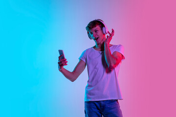 Music in headphones. Young caucasian man's portrait on gradient blue-pink studio background in neon light. Concept of youth, human emotions, facial expression, sales, ad. Beautiful model in casual.