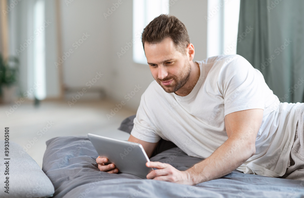 Poster man using tablet lying in bed watching movie online indoors